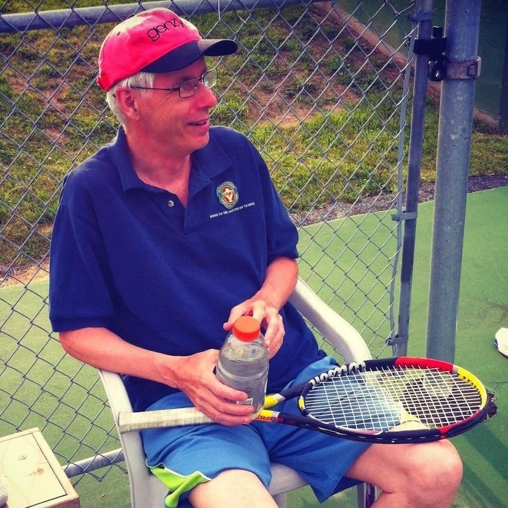 Bruce Cates seated courtside in his pink GenZ cap with a racquet across his lap.