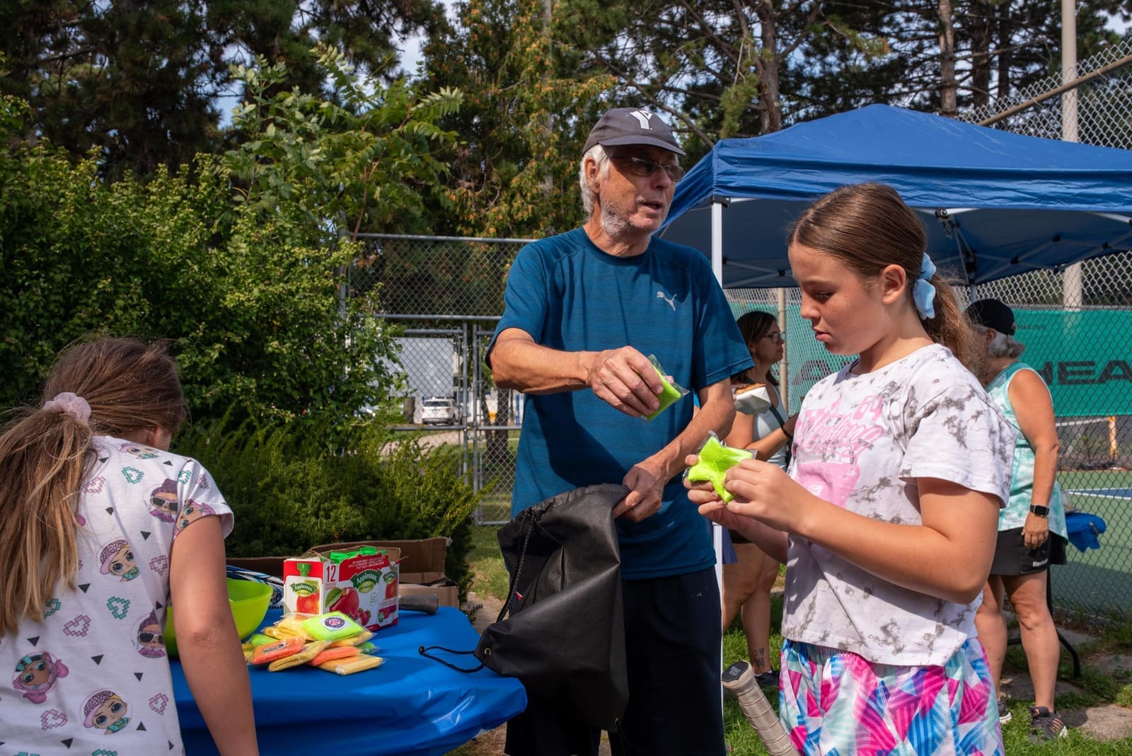 Bruce Cates handing equipment to a young girl at an outdoor tennis event.