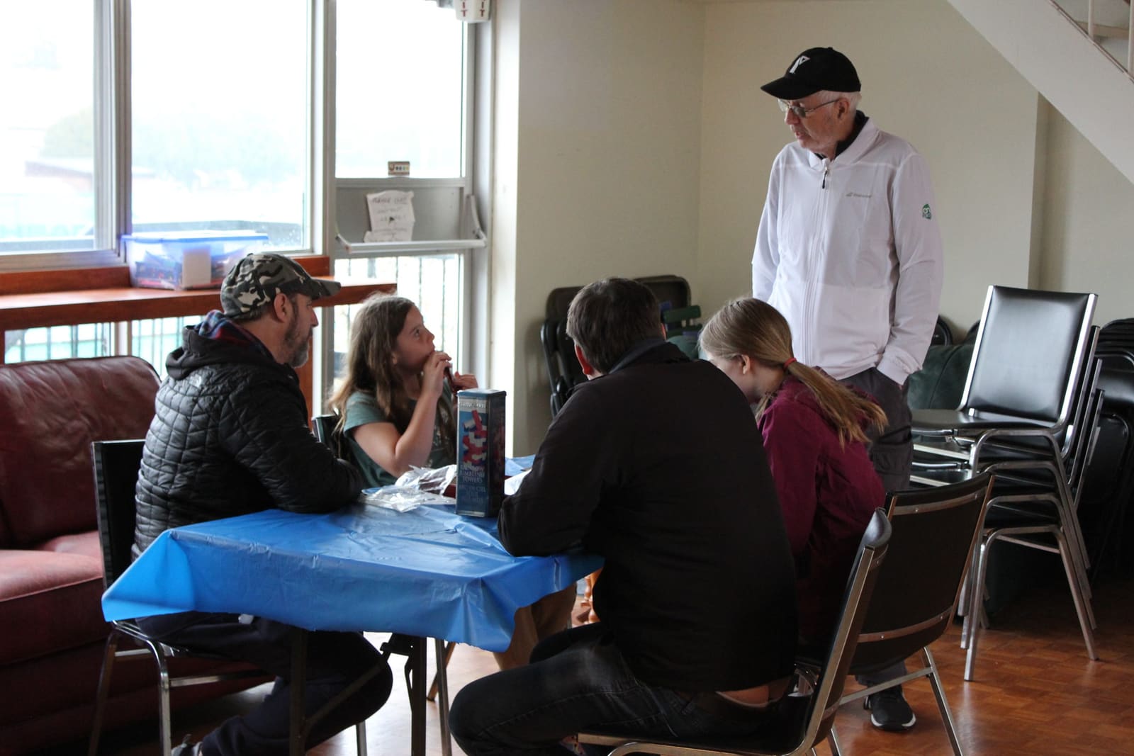 A small indoor gathering at Burlington Tennis Club, with Bruce standing at the head of a covered table while members talk and plan.